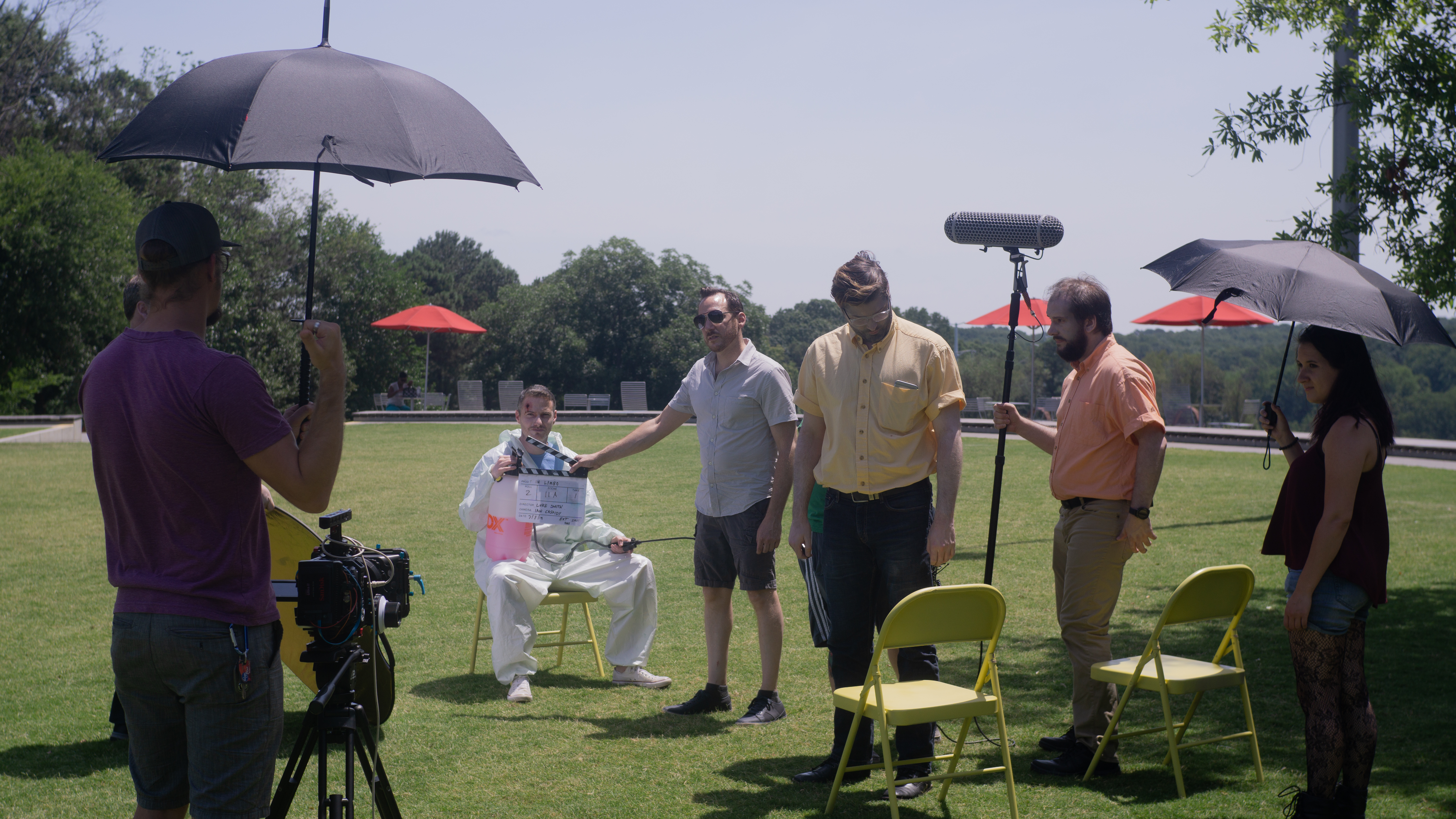 A film crew is preparing to shoot a scene outdoors, with a director holding a clapperboard. The scene features a person in a white suit sitting on a chair, while crew members adjust equipment and hold umbrellas for shade.